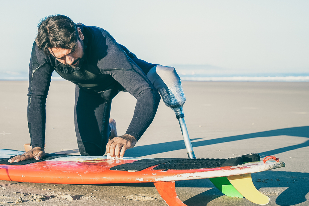 The photo above shows a man waxing his surfboard at the beach. He is an amputee wearing a prosthesis on one leg.