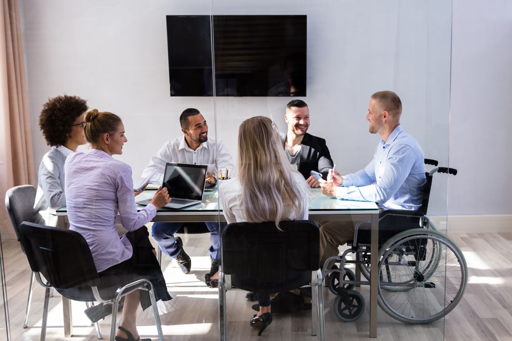 The picture above shows a group of young professionals seated around a table in an office setting engaged in discussion. One member of the team is a white male in a wheelchair.