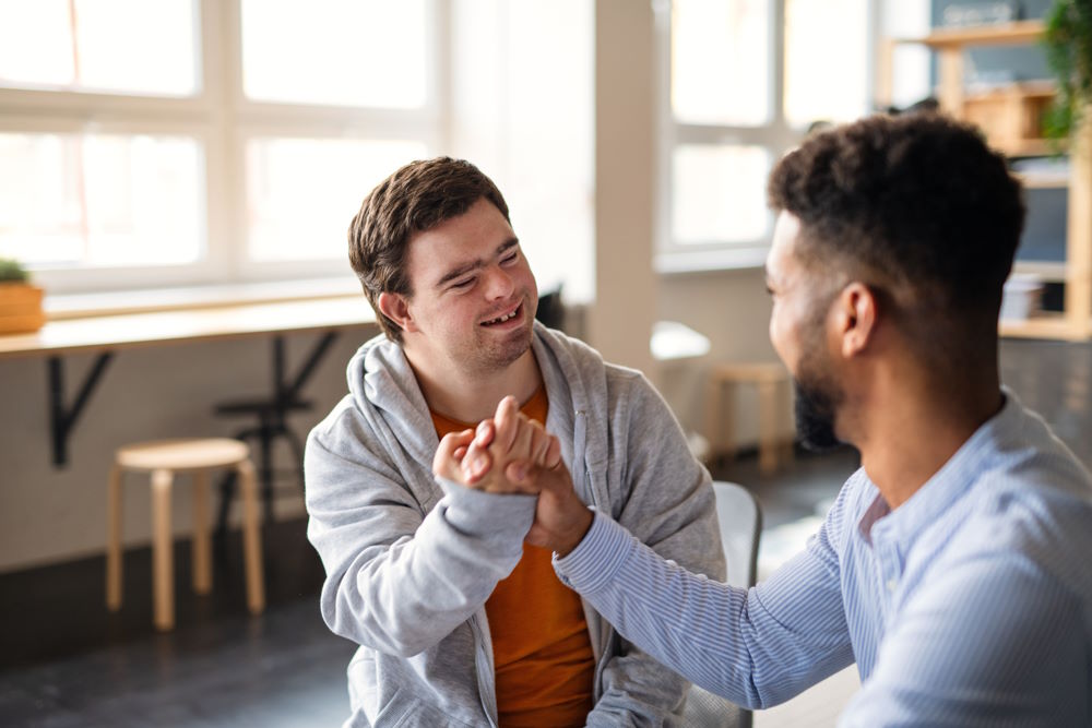 The picture above shows two male friends. They are bonding with a fist bump and smiles. One is a young black male, the other is white who has Down Syndrome.
